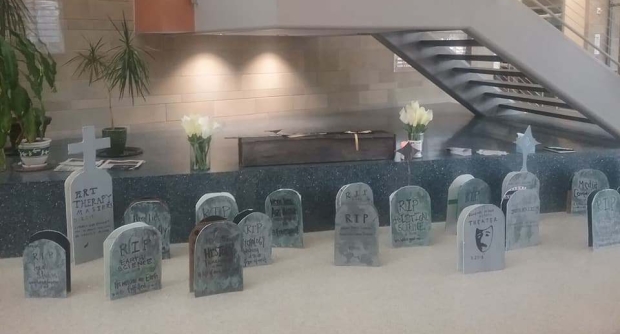 picture is of 25 handmade headstones with the names of academic programs set out in a univeristy building. in the background is a cardboard casket flanked by paper flowers.